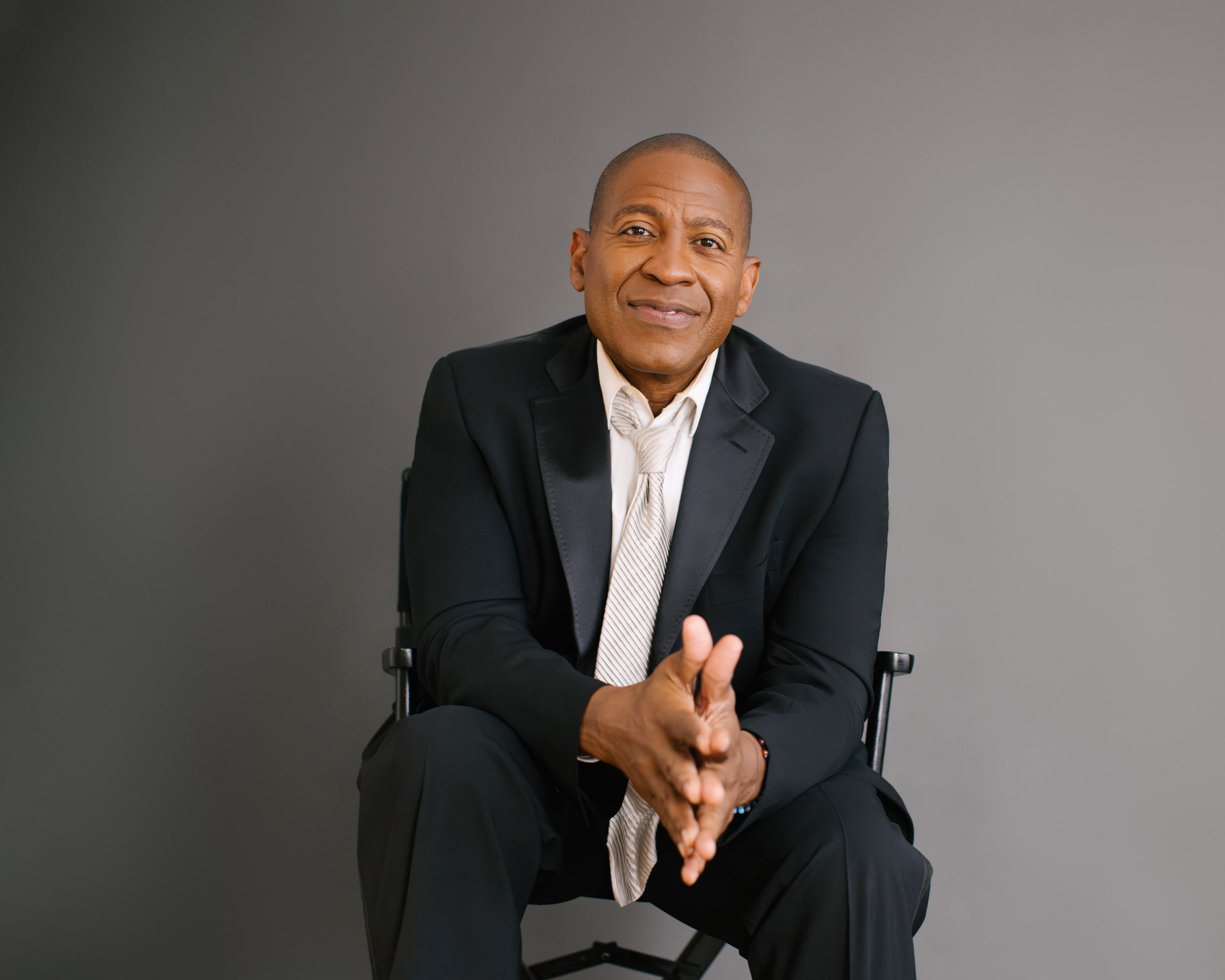 Carlos Watson seated in a black suit, hands clasped, smiling